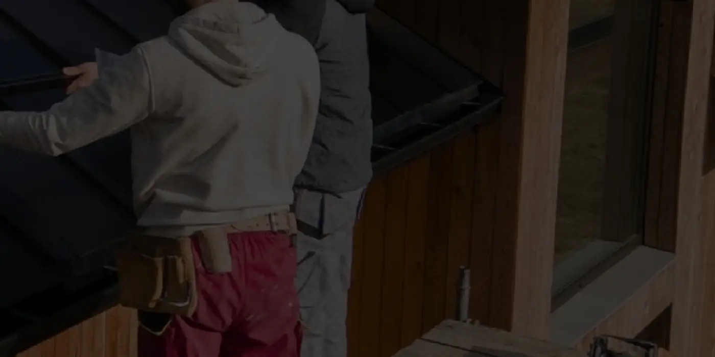 Two roofers working along the eave of a dark metal roof beside timber cladding.